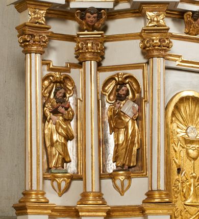 Zdjęcie nr 6: Tabernakulum architektoniczne, jednokondygnacyjne, pięcioosiowe. Osie wydzielone są sześcioma kanelowanymi kolumienkami na wysokich, profilowanych cokołach, z liściastymi kapitelami, podtrzymującymi profilowane belkowanie wyłamane na ich osiach. W polach pomiędzy kolumnami znajdują się nisze ujęte profilowaną ramą z uszakami. W czterech bocznych niszach, na trójdzielnych, profilowanych wspornikach stoją płaskorzeźbione postacie czterech ewangelistów na tle muszli palmetowych. Figurki są całopostaciowe, w ujęciu frontalnym, w złotych szatach, podtrzymują otwarte księgi, wskazując na tekst. Drzwiczki znajdują się na osi środkowej, zamknięte są półkoliście, płaskorzeźbione z przedstawieniem kielicha z koroną cierniową i z hostią w glorii promienistej. Kielich flankowany jest kłosami zbóż i winną latoroślą oraz winogronami. W partii belkowania płaskorzeźbiona dekoracja złożona z naprzemiennie ułożonych główek anielskich o złotych skrzydłach i płaskich rozetek. Tron wystawienia w formie baldachimu, z lambrekinem i chwostami, zwieńczony jest kulą z krzyżem, z podwieszonym z tyłu paludamentem podtrzymywanym przez parę puttów. Struktura polichromowana na biało, profile i detale pozłocone. 