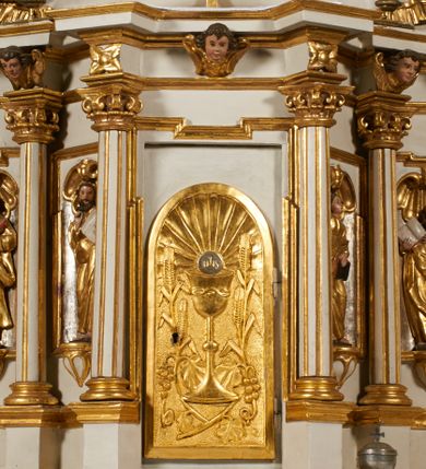 Zdjęcie nr 1: Tabernakulum architektoniczne, jednokondygnacyjne, pięcioosiowe. Osie wydzielone są sześcioma kanelowanymi kolumienkami na wysokich, profilowanych cokołach, z liściastymi kapitelami, podtrzymującymi profilowane belkowanie wyłamane na ich osiach. W polach pomiędzy kolumnami znajdują się nisze ujęte profilowaną ramą z uszakami. W czterech bocznych niszach, na trójdzielnych, profilowanych wspornikach stoją płaskorzeźbione postacie czterech ewangelistów na tle muszli palmetowych. Figurki są całopostaciowe, w ujęciu frontalnym, w złotych szatach, podtrzymują otwarte księgi, wskazując na tekst. Drzwiczki znajdują się na osi środkowej, zamknięte są półkoliście, płaskorzeźbione z przedstawieniem kielicha z koroną cierniową i z hostią w glorii promienistej. Kielich flankowany jest kłosami zbóż i winną latoroślą oraz winogronami. W partii belkowania płaskorzeźbiona dekoracja złożona z naprzemiennie ułożonych główek anielskich o złotych skrzydłach i płaskich rozetek. Tron wystawienia w formie baldachimu, z lambrekinem i chwostami, zwieńczony jest kulą z krzyżem, z podwieszonym z tyłu paludamentem podtrzymywanym przez parę puttów. Struktura polichromowana na biało, profile i detale pozłocone. 
