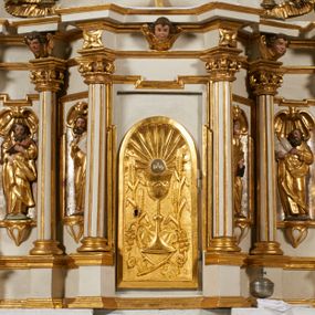 Zdjęcie nr 1: Tabernakulum architektoniczne, jednokondygnacyjne, pięcioosiowe. Osie wydzielone są sześcioma kanelowanymi kolumienkami na wysokich, profilowanych cokołach, z liściastymi kapitelami, podtrzymującymi profilowane belkowanie wyłamane na ich osiach. W polach pomiędzy kolumnami znajdują się nisze ujęte profilowaną ramą z uszakami. W czterech bocznych niszach, na trójdzielnych, profilowanych wspornikach stoją płaskorzeźbione postacie czterech ewangelistów na tle muszli palmetowych. Figurki są całopostaciowe, w ujęciu frontalnym, w złotych szatach, podtrzymują otwarte księgi, wskazując na tekst. Drzwiczki znajdują się na osi środkowej, zamknięte są półkoliście, płaskorzeźbione z przedstawieniem kielicha z koroną cierniową i z hostią w glorii promienistej. Kielich flankowany jest kłosami zbóż i winną latoroślą oraz winogronami. W partii belkowania płaskorzeźbiona dekoracja złożona z naprzemiennie ułożonych główek anielskich o złotych skrzydłach i płaskich rozetek. Tron wystawienia w formie baldachimu, z lambrekinem i chwostami, zwieńczony jest kulą z krzyżem, z podwieszonym z tyłu paludamentem podtrzymywanym przez parę puttów. Struktura polichromowana na biało, profile i detale pozłocone. 