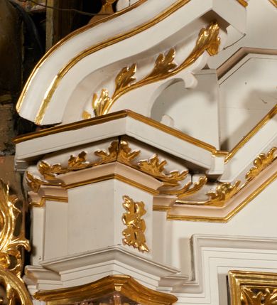 Zdjęcie nr 28: Nastawa architektoniczna, jednoosiowa, dwukondygnacyjna, ustawiona na niewielkim cokole, z dwoma prostopadłościennymi postumentami wydzielonymi po bokach. Ołtarz drewniany, prostopadłościenny, z drewnianą mensą. Antepedium w formie poziomego prostokąta ujętego profilowaną ramą, w polu malowany monogram IHS flankowany gałązkami lilii, u dołu girlanda kwiatowa. W predelli nastawy płycina w formie poziomego prostokąta poszerzonego na krótszych bokach za pomocą nadwieszonych łuków półkolistych, ze sceną modlitwy Chrystusa w Ogrójcu. Pierwsza kondygnacja ujęta parą kolumn stojących na postumentach, o skręconych trzonach dekorowanych ornamentem roślinnym. Podpory dźwigają belkowanie, o pełnych odcinkach nad kolumnami ustawionych pod kątem, z architrawem wyłamującym się schodkowo nad polem środkowym oraz gzymsem przyjmującym formę trzech boków z ośmioboku oraz przerwany, falisty przyczółek. Fryz belkowania ozdobiony jest ornamentem roślinnym, gzyms zaś dekorowany muszlą i zwisami kwiatowymi. Całe pole środkowe wypełnia nisza w kształcie stojącego prostokąta z wciętymi narożami, ujęta ramą ozdobioną ornamentem roślinnym, wewnątrz znajduje się rzeźbiarska grupa Ukrzyżowania na tle malowanego pejzażu. Pierwsza kondygnacja ujęta jest uszami utworzonymi z suchego akantu, w ich dolnej części postumenty dla figur św. Walentego i św. Jana Ewangelisty. Druga kondygnacja jest węższa od poprzedniej, flankowana parą ustawionych lekko pod kątem pilastrów dźwigających gzyms, dekorowanych w partii trzonów kampanulami. W polu obraz ze św. Wawrzyńcem w formie pionowego owalu w profilowanej ramie. Całość zwieńczona okiem Opatrzności na tle obłoków w glorii promienistej. Na belkowaniu niewielkie figury aniołów. Druga kondygnacja ujęta jest pionowymi rzędami kampanul. Struktura polichromowana w kolorze białym, z pozłacanymi profilami i elementami ornamentyki oraz srebrzonymi detalami. 