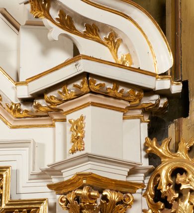 Zdjęcie nr 29: Nastawa architektoniczna, jednoosiowa, dwukondygnacyjna, ustawiona na niewielkim cokole, z dwoma prostopadłościennymi postumentami wydzielonymi po bokach. Ołtarz drewniany, prostopadłościenny, z drewnianą mensą. Antepedium w formie poziomego prostokąta ujętego profilowaną ramą, w polu malowany monogram IHS flankowany gałązkami lilii, u dołu girlanda kwiatowa. W predelli nastawy płycina w formie poziomego prostokąta poszerzonego na krótszych bokach za pomocą nadwieszonych łuków półkolistych, ze sceną modlitwy Chrystusa w Ogrójcu. Pierwsza kondygnacja ujęta parą kolumn stojących na postumentach, o skręconych trzonach dekorowanych ornamentem roślinnym. Podpory dźwigają belkowanie, o pełnych odcinkach nad kolumnami ustawionych pod kątem, z architrawem wyłamującym się schodkowo nad polem środkowym oraz gzymsem przyjmującym formę trzech boków z ośmioboku oraz przerwany, falisty przyczółek. Fryz belkowania ozdobiony jest ornamentem roślinnym, gzyms zaś dekorowany muszlą i zwisami kwiatowymi. Całe pole środkowe wypełnia nisza w kształcie stojącego prostokąta z wciętymi narożami, ujęta ramą ozdobioną ornamentem roślinnym, wewnątrz znajduje się rzeźbiarska grupa Ukrzyżowania na tle malowanego pejzażu. Pierwsza kondygnacja ujęta jest uszami utworzonymi z suchego akantu, w ich dolnej części postumenty dla figur św. Walentego i św. Jana Ewangelisty. Druga kondygnacja jest węższa od poprzedniej, flankowana parą ustawionych lekko pod kątem pilastrów dźwigających gzyms, dekorowanych w partii trzonów kampanulami. W polu obraz ze św. Wawrzyńcem w formie pionowego owalu w profilowanej ramie. Całość zwieńczona okiem Opatrzności na tle obłoków w glorii promienistej. Na belkowaniu niewielkie figury aniołów. Druga kondygnacja ujęta jest pionowymi rzędami kampanul. Struktura polichromowana w kolorze białym, z pozłacanymi profilami i elementami ornamentyki oraz srebrzonymi detalami. 