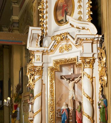 Zdjęcie nr 23: Nastawa architektoniczna, jednoosiowa, dwukondygnacyjna, ustawiona na niewielkim cokole, z dwoma prostopadłościennymi postumentami wydzielonymi po bokach. Ołtarz drewniany, prostopadłościenny, z drewnianą mensą. Antepedium w formie poziomego prostokąta ujętego profilowaną ramą, w polu malowany monogram IHS flankowany gałązkami lilii, u dołu girlanda kwiatowa. W predelli nastawy płycina w formie poziomego prostokąta poszerzonego na krótszych bokach za pomocą nadwieszonych łuków półkolistych, ze sceną modlitwy Chrystusa w Ogrójcu. Pierwsza kondygnacja ujęta parą kolumn stojących na postumentach, o skręconych trzonach dekorowanych ornamentem roślinnym. Podpory dźwigają belkowanie, o pełnych odcinkach nad kolumnami ustawionych pod kątem, z architrawem wyłamującym się schodkowo nad polem środkowym oraz gzymsem przyjmującym formę trzech boków z ośmioboku oraz przerwany, falisty przyczółek. Fryz belkowania ozdobiony jest ornamentem roślinnym, gzyms zaś dekorowany muszlą i zwisami kwiatowymi. Całe pole środkowe wypełnia nisza w kształcie stojącego prostokąta z wciętymi narożami, ujęta ramą ozdobioną ornamentem roślinnym, wewnątrz znajduje się rzeźbiarska grupa Ukrzyżowania na tle malowanego pejzażu. Pierwsza kondygnacja ujęta jest uszami utworzonymi z suchego akantu, w ich dolnej części postumenty dla figur św. Walentego i św. Jana Ewangelisty. Druga kondygnacja jest węższa od poprzedniej, flankowana parą ustawionych lekko pod kątem pilastrów dźwigających gzyms, dekorowanych w partii trzonów kampanulami. W polu obraz ze św. Wawrzyńcem w formie pionowego owalu w profilowanej ramie. Całość zwieńczona okiem Opatrzności na tle obłoków w glorii promienistej. Na belkowaniu niewielkie figury aniołów. Druga kondygnacja ujęta jest pionowymi rzędami kampanul. Struktura polichromowana w kolorze białym, z pozłacanymi profilami i elementami ornamentyki oraz srebrzonymi detalami. 