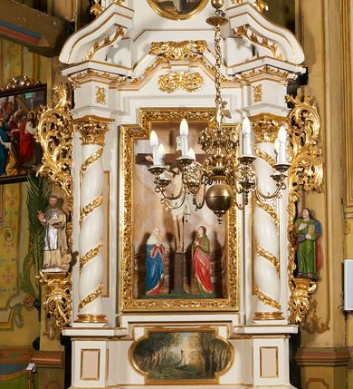 Zdjęcie nr 13: Nastawa architektoniczna, jednoosiowa, dwukondygnacyjna, ustawiona na niewielkim cokole, z dwoma prostopadłościennymi postumentami wydzielonymi po bokach. Ołtarz drewniany, prostopadłościenny, z drewnianą mensą. Antepedium w formie poziomego prostokąta ujętego profilowaną ramą, w polu malowany monogram IHS flankowany gałązkami lilii, u dołu girlanda kwiatowa. W predelli nastawy płycina w formie poziomego prostokąta poszerzonego na krótszych bokach za pomocą nadwieszonych łuków półkolistych, ze sceną modlitwy Chrystusa w Ogrójcu. Pierwsza kondygnacja ujęta parą kolumn stojących na postumentach, o skręconych trzonach dekorowanych ornamentem roślinnym. Podpory dźwigają belkowanie, o pełnych odcinkach nad kolumnami ustawionych pod kątem, z architrawem wyłamującym się schodkowo nad polem środkowym oraz gzymsem przyjmującym formę trzech boków z ośmioboku oraz przerwany, falisty przyczółek. Fryz belkowania ozdobiony jest ornamentem roślinnym, gzyms zaś dekorowany muszlą i zwisami kwiatowymi. Całe pole środkowe wypełnia nisza w kształcie stojącego prostokąta z wciętymi narożami, ujęta ramą ozdobioną ornamentem roślinnym, wewnątrz znajduje się rzeźbiarska grupa Ukrzyżowania na tle malowanego pejzażu. Pierwsza kondygnacja ujęta jest uszami utworzonymi z suchego akantu, w ich dolnej części postumenty dla figur św. Walentego i św. Jana Ewangelisty. Druga kondygnacja jest węższa od poprzedniej, flankowana parą ustawionych lekko pod kątem pilastrów dźwigających gzyms, dekorowanych w partii trzonów kampanulami. W polu obraz ze św. Wawrzyńcem w formie pionowego owalu w profilowanej ramie. Całość zwieńczona okiem Opatrzności na tle obłoków w glorii promienistej. Na belkowaniu niewielkie figury aniołów. Druga kondygnacja ujęta jest pionowymi rzędami kampanul. Struktura polichromowana w kolorze białym, z pozłacanymi profilami i elementami ornamentyki oraz srebrzonymi detalami. 