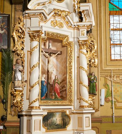 Zdjęcie nr 2: Nastawa architektoniczna, jednoosiowa, dwukondygnacyjna, ustawiona na niewielkim cokole, z dwoma prostopadłościennymi postumentami wydzielonymi po bokach. Ołtarz drewniany, prostopadłościenny, z drewnianą mensą. Antepedium w formie poziomego prostokąta ujętego profilowaną ramą, w polu malowany monogram IHS flankowany gałązkami lilii, u dołu girlanda kwiatowa. W predelli nastawy płycina w formie poziomego prostokąta poszerzonego na krótszych bokach za pomocą nadwieszonych łuków półkolistych, ze sceną modlitwy Chrystusa w Ogrójcu. Pierwsza kondygnacja ujęta parą kolumn stojących na postumentach, o skręconych trzonach dekorowanych ornamentem roślinnym. Podpory dźwigają belkowanie, o pełnych odcinkach nad kolumnami ustawionych pod kątem, z architrawem wyłamującym się schodkowo nad polem środkowym oraz gzymsem przyjmującym formę trzech boków z ośmioboku oraz przerwany, falisty przyczółek. Fryz belkowania ozdobiony jest ornamentem roślinnym, gzyms zaś dekorowany muszlą i zwisami kwiatowymi. Całe pole środkowe wypełnia nisza w kształcie stojącego prostokąta z wciętymi narożami, ujęta ramą ozdobioną ornamentem roślinnym, wewnątrz znajduje się rzeźbiarska grupa Ukrzyżowania na tle malowanego pejzażu. Pierwsza kondygnacja ujęta jest uszami utworzonymi z suchego akantu, w ich dolnej części postumenty dla figur św. Walentego i św. Jana Ewangelisty. Druga kondygnacja jest węższa od poprzedniej, flankowana parą ustawionych lekko pod kątem pilastrów dźwigających gzyms, dekorowanych w partii trzonów kampanulami. W polu obraz ze św. Wawrzyńcem w formie pionowego owalu w profilowanej ramie. Całość zwieńczona okiem Opatrzności na tle obłoków w glorii promienistej. Na belkowaniu niewielkie figury aniołów. Druga kondygnacja ujęta jest pionowymi rzędami kampanul. Struktura polichromowana w kolorze białym, z pozłacanymi profilami i elementami ornamentyki oraz srebrzonymi detalami. 