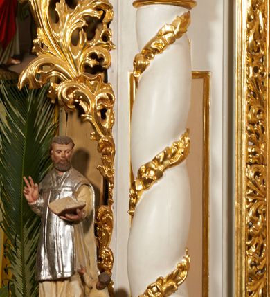 Zdjęcie nr 25: Nastawa architektoniczna, jednoosiowa, dwukondygnacyjna, ustawiona na niewielkim cokole, z dwoma prostopadłościennymi postumentami wydzielonymi po bokach. Ołtarz drewniany, prostopadłościenny, z drewnianą mensą. Antepedium w formie poziomego prostokąta ujętego profilowaną ramą, w polu malowany monogram IHS flankowany gałązkami lilii, u dołu girlanda kwiatowa. W predelli nastawy płycina w formie poziomego prostokąta poszerzonego na krótszych bokach za pomocą nadwieszonych łuków półkolistych, ze sceną modlitwy Chrystusa w Ogrójcu. Pierwsza kondygnacja ujęta parą kolumn stojących na postumentach, o skręconych trzonach dekorowanych ornamentem roślinnym. Podpory dźwigają belkowanie, o pełnych odcinkach nad kolumnami ustawionych pod kątem, z architrawem wyłamującym się schodkowo nad polem środkowym oraz gzymsem przyjmującym formę trzech boków z ośmioboku oraz przerwany, falisty przyczółek. Fryz belkowania ozdobiony jest ornamentem roślinnym, gzyms zaś dekorowany muszlą i zwisami kwiatowymi. Całe pole środkowe wypełnia nisza w kształcie stojącego prostokąta z wciętymi narożami, ujęta ramą ozdobioną ornamentem roślinnym, wewnątrz znajduje się rzeźbiarska grupa Ukrzyżowania na tle malowanego pejzażu. Pierwsza kondygnacja ujęta jest uszami utworzonymi z suchego akantu, w ich dolnej części postumenty dla figur św. Walentego i św. Jana Ewangelisty. Druga kondygnacja jest węższa od poprzedniej, flankowana parą ustawionych lekko pod kątem pilastrów dźwigających gzyms, dekorowanych w partii trzonów kampanulami. W polu obraz ze św. Wawrzyńcem w formie pionowego owalu w profilowanej ramie. Całość zwieńczona okiem Opatrzności na tle obłoków w glorii promienistej. Na belkowaniu niewielkie figury aniołów. Druga kondygnacja ujęta jest pionowymi rzędami kampanul. Struktura polichromowana w kolorze białym, z pozłacanymi profilami i elementami ornamentyki oraz srebrzonymi detalami. 