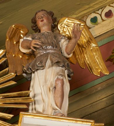 Zdjęcie nr 19: Nastawa architektoniczna, jednoosiowa, dwukondygnacyjna, ustawiona na niewielkim cokole, z dwoma prostopadłościennymi postumentami wydzielonymi po bokach. Ołtarz drewniany, prostopadłościenny, z drewnianą mensą. Antepedium w formie poziomego prostokąta ujętego profilowaną ramą, w polu malowany monogram IHS flankowany gałązkami lilii, u dołu girlanda kwiatowa. W predelli nastawy płycina w formie poziomego prostokąta poszerzonego na krótszych bokach za pomocą nadwieszonych łuków półkolistych, ze sceną modlitwy Chrystusa w Ogrójcu. Pierwsza kondygnacja ujęta parą kolumn stojących na postumentach, o skręconych trzonach dekorowanych ornamentem roślinnym. Podpory dźwigają belkowanie, o pełnych odcinkach nad kolumnami ustawionych pod kątem, z architrawem wyłamującym się schodkowo nad polem środkowym oraz gzymsem przyjmującym formę trzech boków z ośmioboku oraz przerwany, falisty przyczółek. Fryz belkowania ozdobiony jest ornamentem roślinnym, gzyms zaś dekorowany muszlą i zwisami kwiatowymi. Całe pole środkowe wypełnia nisza w kształcie stojącego prostokąta z wciętymi narożami, ujęta ramą ozdobioną ornamentem roślinnym, wewnątrz znajduje się rzeźbiarska grupa Ukrzyżowania na tle malowanego pejzażu. Pierwsza kondygnacja ujęta jest uszami utworzonymi z suchego akantu, w ich dolnej części postumenty dla figur św. Walentego i św. Jana Ewangelisty. Druga kondygnacja jest węższa od poprzedniej, flankowana parą ustawionych lekko pod kątem pilastrów dźwigających gzyms, dekorowanych w partii trzonów kampanulami. W polu obraz ze św. Wawrzyńcem w formie pionowego owalu w profilowanej ramie. Całość zwieńczona okiem Opatrzności na tle obłoków w glorii promienistej. Na belkowaniu niewielkie figury aniołów. Druga kondygnacja ujęta jest pionowymi rzędami kampanul. Struktura polichromowana w kolorze białym, z pozłacanymi profilami i elementami ornamentyki oraz srebrzonymi detalami. 