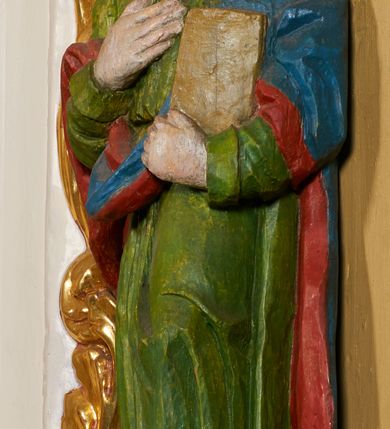 Zdjęcie nr 1: Rzeźba pełna, święty ustawiony jest frontalnie, w kontrapoście, z głową skierowaną w prawo, lewą ręką przyciska do boku księgę, prawą kładzie na piersi. Twarz świętego jest okrągła o dużych oczach skierowanych w bok, prostym nosie i pełnych ustach, okolona długimi, falowanymi włosami opadającymi na ramiona i plecy. Ubrany jest w zieloną suknię, przepasaną w talii i niebiesko-czerwony płaszcz zarzucony na lewe ramię, stopy świętego bose. Polichromia ciała traktowana naturalistycznie.