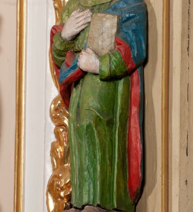 Zdjęcie nr 4: Rzeźba pełna, święty ustawiony jest frontalnie, w kontrapoście, z głową skierowaną w prawo, lewą ręką przyciska do boku księgę, prawą kładzie na piersi. Twarz świętego jest okrągła o dużych oczach skierowanych w bok, prostym nosie i pełnych ustach, okolona długimi, falowanymi włosami opadającymi na ramiona i plecy. Ubrany jest w zieloną suknię, przepasaną w talii i niebiesko-czerwony płaszcz zarzucony na lewe ramię, stopy świętego bose. Polichromia ciała traktowana naturalistycznie.