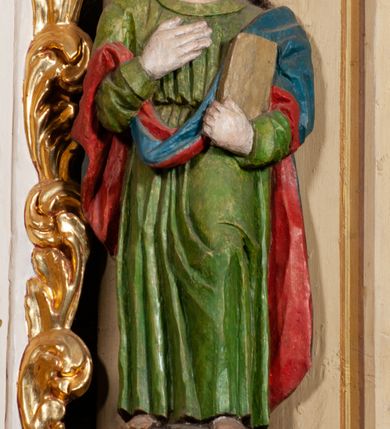 Zdjęcie nr 3: Rzeźba pełna, święty ustawiony jest frontalnie, w kontrapoście, z głową skierowaną w prawo, lewą ręką przyciska do boku księgę, prawą kładzie na piersi. Twarz świętego jest okrągła o dużych oczach skierowanych w bok, prostym nosie i pełnych ustach, okolona długimi, falowanymi włosami opadającymi na ramiona i plecy. Ubrany jest w zieloną suknię, przepasaną w talii i niebiesko-czerwony płaszcz zarzucony na lewe ramię, stopy świętego bose. Polichromia ciała traktowana naturalistycznie.