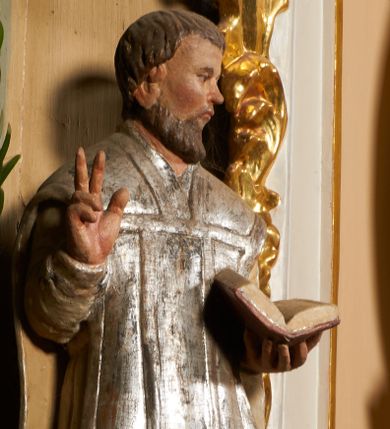 Zdjęcie nr 1: Święty został ukazany frontalnie, w wykroku, z głową skierowaną w lewo, w wykroku, lewą ręką podtrzymuje otwartą księgę, prawą unosi w geście błogosławieństwa. Jego twarz jest pociągła, o małych oczach, krótkim nosie i wąskich ustach, okolona brodą i krótkimi włosami. Ubrany jest w białą albę i posrebrzany ornat. Przy jego lewej nodze półpostać mężczyzny, który prawą ręką trzyma szatę świętego, ubrany jest w białą, przepasaną w talii tunikę. Polichromia ciała naturalistyczna, ornat posrebrzany.

