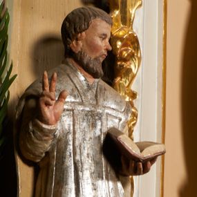 Zdjęcie nr 1: Święty został ukazany frontalnie, w wykroku, z głową skierowaną w lewo, w wykroku, lewą ręką podtrzymuje otwartą księgę, prawą unosi w geście błogosławieństwa. Jego twarz jest pociągła, o małych oczach, krótkim nosie i wąskich ustach, okolona brodą i krótkimi włosami. Ubrany jest w białą albę i posrebrzany ornat. Przy jego lewej nodze półpostać mężczyzny, który prawą ręką trzyma szatę świętego, ubrany jest w białą, przepasaną w talii tunikę. Polichromia ciała naturalistyczna, ornat posrebrzany.

