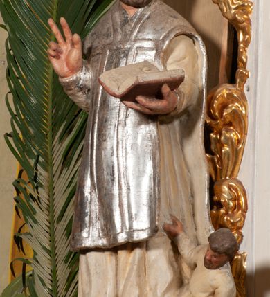 Zdjęcie nr 2: Święty został ukazany frontalnie, w wykroku, z głową skierowaną w lewo, w wykroku, lewą ręką podtrzymuje otwartą księgę, prawą unosi w geście błogosławieństwa. Jego twarz jest pociągła, o małych oczach, krótkim nosie i wąskich ustach, okolona brodą i krótkimi włosami. Ubrany jest w białą albę i posrebrzany ornat. Przy jego lewej nodze półpostać mężczyzny, który prawą ręką trzyma szatę świętego, ubrany jest w białą, przepasaną w talii tunikę. Polichromia ciała naturalistyczna, ornat posrebrzany.

