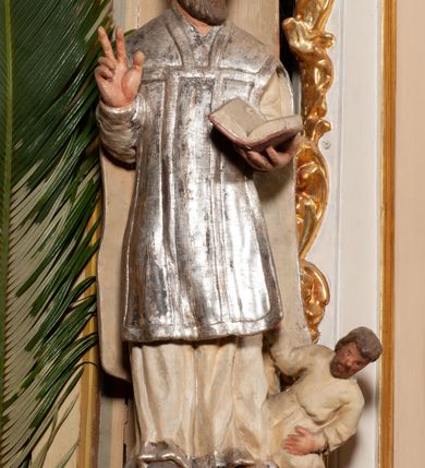 Zdjęcie nr 3: Święty został ukazany frontalnie, w wykroku, z głową skierowaną w lewo, w wykroku, lewą ręką podtrzymuje otwartą księgę, prawą unosi w geście błogosławieństwa. Jego twarz jest pociągła, o małych oczach, krótkim nosie i wąskich ustach, okolona brodą i krótkimi włosami. Ubrany jest w białą albę i posrebrzany ornat. Przy jego lewej nodze półpostać mężczyzny, który prawą ręką trzyma szatę świętego, ubrany jest w białą, przepasaną w talii tunikę. Polichromia ciała naturalistyczna, ornat posrebrzany.

