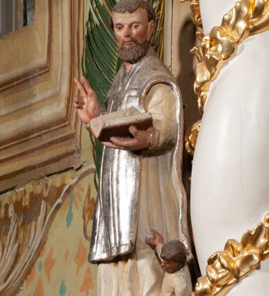 Zdjęcie nr 4: Święty został ukazany frontalnie, w wykroku, z głową skierowaną w lewo, w wykroku, lewą ręką podtrzymuje otwartą księgę, prawą unosi w geście błogosławieństwa. Jego twarz jest pociągła, o małych oczach, krótkim nosie i wąskich ustach, okolona brodą i krótkimi włosami. Ubrany jest w białą albę i posrebrzany ornat. Przy jego lewej nodze półpostać mężczyzny, który prawą ręką trzyma szatę świętego, ubrany jest w białą, przepasaną w talii tunikę. Polichromia ciała naturalistyczna, ornat posrebrzany.

