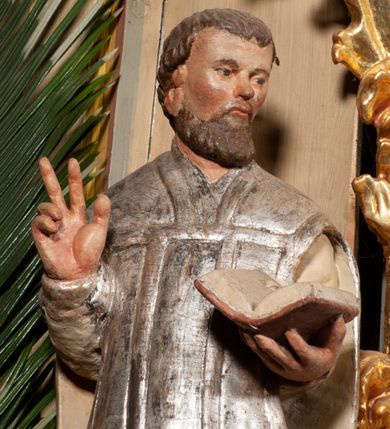 Zdjęcie nr 5: Święty został ukazany frontalnie, w wykroku, z głową skierowaną w lewo, w wykroku, lewą ręką podtrzymuje otwartą księgę, prawą unosi w geście błogosławieństwa. Jego twarz jest pociągła, o małych oczach, krótkim nosie i wąskich ustach, okolona brodą i krótkimi włosami. Ubrany jest w białą albę i posrebrzany ornat. Przy jego lewej nodze półpostać mężczyzny, który prawą ręką trzyma szatę świętego, ubrany jest w białą, przepasaną w talii tunikę. Polichromia ciała naturalistyczna, ornat posrebrzany.

