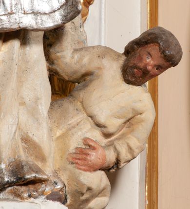 Zdjęcie nr 7: Święty został ukazany frontalnie, w wykroku, z głową skierowaną w lewo, w wykroku, lewą ręką podtrzymuje otwartą księgę, prawą unosi w geście błogosławieństwa. Jego twarz jest pociągła, o małych oczach, krótkim nosie i wąskich ustach, okolona brodą i krótkimi włosami. Ubrany jest w białą albę i posrebrzany ornat. Przy jego lewej nodze półpostać mężczyzny, który prawą ręką trzyma szatę świętego, ubrany jest w białą, przepasaną w talii tunikę. Polichromia ciała naturalistyczna, ornat posrebrzany.

