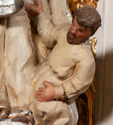 Zdjęcie nr 6: Święty został ukazany frontalnie, w wykroku, z głową skierowaną w lewo, w wykroku, lewą ręką podtrzymuje otwartą księgę, prawą unosi w geście błogosławieństwa. Jego twarz jest pociągła, o małych oczach, krótkim nosie i wąskich ustach, okolona brodą i krótkimi włosami. Ubrany jest w białą albę i posrebrzany ornat. Przy jego lewej nodze półpostać mężczyzny, który prawą ręką trzyma szatę świętego, ubrany jest w białą, przepasaną w talii tunikę. Polichromia ciała naturalistyczna, ornat posrebrzany.

