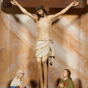 Zdjęcie nr 1: Rzeźbiona grupa Ukrzyżowania znajduje się we wnęce, ujętej ramą, zdobioną stylizowanymi liśćmi, na niewysokim podeście. Umarły Chrystus przybity jest do drzewa krzyża trzema gwoździami, ukazany frontalnie, z głową lekko opadającą na prawe ramię. Ciało w niewielkim zwisie, z ramionami układającymi się w łuk, o delikatnym modelunku mięśni. Chrystus ma pociągłą twarz, którą okalają broda i wąsy. Włosy swobodnie opadają na ramiona. Na głowie korona cierniowa. Jego biodra przepasane są białym perizonium, sięgającym do połowy ud, zdobionym złotą lamówką, zawiązanym na lewym boku ze zwisem tkaniny. Po lewej stronie krzyża stoi Matka Boska, obrócona ku niemu w trzech czwartych, z rękami skrzyżowanymi na piersiach. Twarz ma pociągłą, z zamkniętymi oczami. Maria odziana jest w czerwoną suknię, niebieski płaszcz, zdobiony złotą bordiurą i geometrycznym ornamentem, na głowie ma biały welon, owinięty wokół szyi. Po prawej stronie krzyża znajduje się postać św. Jana Ewangelisty, również zwróconego w trzech czwartych ku krzyżowi, przedstawionego w kontrapoście, z rękami skrzyżowanymi na piersiach. Jego głowa jest lekko uniesiona do góry, twarz jest pociągła. Włosy swobodnie opadające na ramiona. Święty odziany jest w zieloną tunikę, zdobioną złotą lamówką oraz czerwony płaszcz, zarzucony na prawe ramię i podtrzymywany z przodu. Grupa Ukrzyżowania ukazana została na malowanym monochromatycznie tle we wnęce przedstawiającym skalisty pejzaż. Figury w partiach ciała polichromowane są naturalistycznie, detale szat pozłacane. 