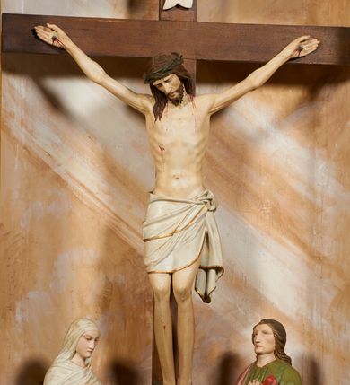 Zdjęcie nr 3: Rzeźbiona grupa Ukrzyżowania znajduje się we wnęce, ujętej ramą, zdobioną stylizowanymi liśćmi, na niewysokim podeście. Umarły Chrystus przybity jest do drzewa krzyża trzema gwoździami, ukazany frontalnie, z głową lekko opadającą na prawe ramię. Ciało w niewielkim zwisie, z ramionami układającymi się w łuk, o delikatnym modelunku mięśni. Chrystus ma pociągłą twarz, którą okalają broda i wąsy. Włosy swobodnie opadają na ramiona. Na głowie korona cierniowa. Jego biodra przepasane są białym perizonium, sięgającym do połowy ud, zdobionym złotą lamówką, zawiązanym na lewym boku ze zwisem tkaniny. Po lewej stronie krzyża stoi Matka Boska, obrócona ku niemu w trzech czwartych, z rękami skrzyżowanymi na piersiach. Twarz ma pociągłą, z zamkniętymi oczami. Maria odziana jest w czerwoną suknię, niebieski płaszcz, zdobiony złotą bordiurą i geometrycznym ornamentem, na głowie ma biały welon, owinięty wokół szyi. Po prawej stronie krzyża znajduje się postać św. Jana Ewangelisty, również zwróconego w trzech czwartych ku krzyżowi, przedstawionego w kontrapoście, z rękami skrzyżowanymi na piersiach. Jego głowa jest lekko uniesiona do góry, twarz jest pociągła. Włosy swobodnie opadające na ramiona. Święty odziany jest w zieloną tunikę, zdobioną złotą lamówką oraz czerwony płaszcz, zarzucony na prawe ramię i podtrzymywany z przodu. Grupa Ukrzyżowania ukazana została na malowanym monochromatycznie tle we wnęce przedstawiającym skalisty pejzaż. Figury w partiach ciała polichromowane są naturalistycznie, detale szat pozłacane. 