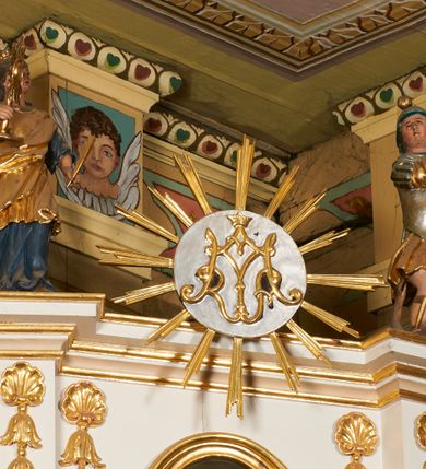 Zdjęcie nr 9: Nastawa ołtarzowa architektoniczna, jednoosiowa, dwukondygnacyjna, ustawiona na niewielkim cokole, z dwoma prostopadłościennymi postumentami wydzielonymi po bokach. Ołtarz drewniany, prostopadłościenny, z drewnianą mensą. Antepedium w formie poziomego prostokąta ujętego profilowaną ramą, w polu malowany monogram Marii, flankowany różanymi gałązkami. W predelli nastawy znajduje się płycina w formie poziomego prostokąta poszerzonego na krótszych bokach za pomocą nadwieszonych łuków półkolistych, ze sceną Zwiastowania. 
Pierwsza kondygnacja ujęta jest parą kolumn stojących na postumentach, o skręconych trzonach dekorowanych ornamentem roślinnym. Podpory dźwigają belkowanie, którego pełne odcinki nad kolumnami ustawione są pod kątem, architraw nad polem środkowym wyłamuje się schodkowo, a gzyms przyjmuje formę trzech boków z ośmioboku. Wyżej widoczny oraz przerwany falisty przyczółek. Fryz belkowania ozdobiony jest palmetami oraz dekoracją roślinną, gzyms dekorowany palmetą. Całe pole środkowe wypełnia obraz Matki Boskiej Różańcowej ze św. Dominikiem i św. Katarzyną Sieneńską w formie pionowego prostokąta z wciętymi narożami, w ramie ozdobionej stylizowanym ornamentem. Pierwsza kondygnacja ujęta jest częściowo ażurowymi uszami utworzonymi z akantu i karbowanej wstęgi. Druga kondygnacja jest węższa, flankowana parą ustawionych lekko pod kątem pilastrów, dekorowanych w partii trzonów kampanulami, dźwigających gzyms,. W polu obraz Objawienia Matki Boskiej w Lourdes w formie pionowego owalu w profilowanej ramie, flankowany kampanulami. Całość zwieńczona medalionem w glorii z monogramem Marii. Na belkowaniu niewielkie figury św. Barbary i św. Michała Archanioła. Druga kondygnacja ujęta jest pionowymi rzędami kampanul. Struktura polichromowana w kolorze białym z pozłacanymi profilami i elementami ornamentyki. 


