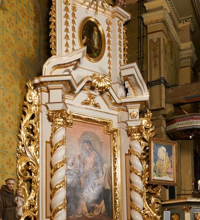 Zdjęcie nr 8: Nastawa ołtarzowa architektoniczna, jednoosiowa, dwukondygnacyjna, ustawiona na niewielkim cokole, z dwoma prostopadłościennymi postumentami wydzielonymi po bokach. Ołtarz drewniany, prostopadłościenny, z drewnianą mensą. Antepedium w formie poziomego prostokąta ujętego profilowaną ramą, w polu malowany monogram Marii, flankowany różanymi gałązkami. W predelli nastawy znajduje się płycina w formie poziomego prostokąta poszerzonego na krótszych bokach za pomocą nadwieszonych łuków półkolistych, ze sceną Zwiastowania. 
Pierwsza kondygnacja ujęta jest parą kolumn stojących na postumentach, o skręconych trzonach dekorowanych ornamentem roślinnym. Podpory dźwigają belkowanie, którego pełne odcinki nad kolumnami ustawione są pod kątem, architraw nad polem środkowym wyłamuje się schodkowo, a gzyms przyjmuje formę trzech boków z ośmioboku. Wyżej widoczny oraz przerwany falisty przyczółek. Fryz belkowania ozdobiony jest palmetami oraz dekoracją roślinną, gzyms dekorowany palmetą. Całe pole środkowe wypełnia obraz Matki Boskiej Różańcowej ze św. Dominikiem i św. Katarzyną Sieneńską w formie pionowego prostokąta z wciętymi narożami, w ramie ozdobionej stylizowanym ornamentem. Pierwsza kondygnacja ujęta jest częściowo ażurowymi uszami utworzonymi z akantu i karbowanej wstęgi. Druga kondygnacja jest węższa, flankowana parą ustawionych lekko pod kątem pilastrów, dekorowanych w partii trzonów kampanulami, dźwigających gzyms,. W polu obraz Objawienia Matki Boskiej w Lourdes w formie pionowego owalu w profilowanej ramie, flankowany kampanulami. Całość zwieńczona medalionem w glorii z monogramem Marii. Na belkowaniu niewielkie figury św. Barbary i św. Michała Archanioła. Druga kondygnacja ujęta jest pionowymi rzędami kampanul. Struktura polichromowana w kolorze białym z pozłacanymi profilami i elementami ornamentyki. 


