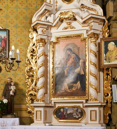 Zdjęcie nr 9: Obraz w formie stojącego prostokąta z wciętymi górnymi narożami z przedstawieniem tronującej Matki Boskiej z Dzieciątkiem wręczających różańce klęczącym przed nimi świętym dominikańskim – św. Dominikowi i św. Katarzynie Sieneńskiej. Maria siedzi na architektoniczno-rzeźbiarskim, ustawionym na dwóch stopniach tronie o renesansowych formach widocznego z tyłu zaplecka. Matka Boska zwrócona jest w trzech czwartych w prawo, lewą ręką podtrzymuje siedzące na jej kolanach Dzieciątko, prawą podaje różaniec św. Dominikowi. Ubrana jest w czerwoną suknię oraz niebieski płaszcz opadający na ramiona i kolana, z kapturem założonym na głowę, wokół której widnieje wieniec z gwiazd. Dzieciątko przechylone jest w lewo i podaje różaniec św. Katarzynie, przepasane jest w partii bioder białą tkaniną. Święci klęczą poniżej, ukazani są w trzech czwartych, zwróceni ku postaci Marii, tyłem do widza. Święty Dominik z dłońmi wyciągniętymi do przodu, św. Katarzyna ze złożonymi na piersi. Ubrani są w habity dominikańskie, św. Katarzyna przedstawiona ze swoim atrybutem – koroną cierniową na głowie. Scena ukazana na tle różowo-błękitnego nieba, wśród obłoków. W prawym dolnym rogu sygnatura „L[eonard] Stroynowski 1921”. Obraz ujęty ramą w formie stojącego prostokąta z wciętymi górnymi narożami dekorowaną rzeźbionym i ażurowym ornamentem utworzonym z cęgów i wolut.
