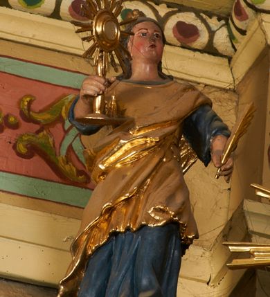 Zdjęcie nr 2: Postać ukazana jest frontalnie, z głową skierowaną delikatnie w lewo, w lewej ręce trzyma liść palmy, w prawej monstrancję. Święta ma okrągłą twarz o pełnych policzkach, okoloną długimi włosami spiętymi z tyłu głowy i opadającymi na plecy. Ubrana jest w długą, niebieską suknię spodnią, pozłacaną suknię wierzchnią sięgającą kolan, przepasaną w talii oraz pozłacany płaszcz przewiązany diagonalnie przez lewe ramię. Na głowie ma koronę. Polichromia naturalistyczna, część szat i atrybuty pozłacane.
