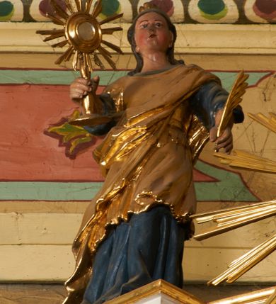 Zdjęcie nr 1: Postać ukazana jest frontalnie, z głową skierowaną delikatnie w lewo, w lewej ręce trzyma liść palmy, w prawej monstrancję. Święta ma okrągłą twarz o pełnych policzkach, okoloną długimi włosami spiętymi z tyłu głowy i opadającymi na plecy. Ubrana jest w długą, niebieską suknię spodnią, pozłacaną suknię wierzchnią sięgającą kolan, przepasaną w talii oraz pozłacany płaszcz przewiązany diagonalnie przez lewe ramię. Na głowie ma koronę. Polichromia naturalistyczna, część szat i atrybuty pozłacane.
