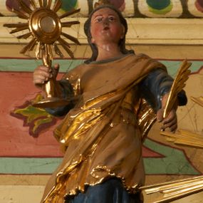 Zdjęcie nr 1: Postać ukazana jest frontalnie, z głową skierowaną delikatnie w lewo, w lewej ręce trzyma liść palmy, w prawej monstrancję. Święta ma okrągłą twarz o pełnych policzkach, okoloną długimi włosami spiętymi z tyłu głowy i opadającymi na plecy. Ubrana jest w długą, niebieską suknię spodnią, pozłacaną suknię wierzchnią sięgającą kolan, przepasaną w talii oraz pozłacany płaszcz przewiązany diagonalnie przez lewe ramię. Na głowie ma koronę. Polichromia naturalistyczna, część szat i atrybuty pozłacane.
