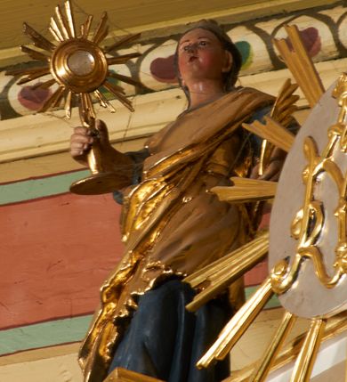 Zdjęcie nr 5: Postać ukazana jest frontalnie, z głową skierowaną delikatnie w lewo, w lewej ręce trzyma liść palmy, w prawej monstrancję. Święta ma okrągłą twarz o pełnych policzkach, okoloną długimi włosami spiętymi z tyłu głowy i opadającymi na plecy. Ubrana jest w długą, niebieską suknię spodnią, pozłacaną suknię wierzchnią sięgającą kolan, przepasaną w talii oraz pozłacany płaszcz przewiązany diagonalnie przez lewe ramię. Na głowie ma koronę. Polichromia naturalistyczna, część szat i atrybuty pozłacane.
