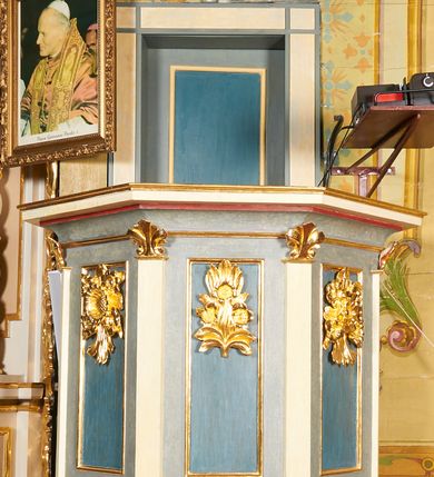 Zdjęcie nr 2: Ambona jest przyścienna, wisząca, z dostępem od zakrystii przez drzwiczki w zaplecku. Kosz ma na planie połowy ośmioboku, na podstawie w formie wałka dekorowanego liśćmi, z podwieszeniem zakończonym szyszką. Ścianki kosza wydzielone są pilastrami rozmieszczonymi w narożach wieloboku, zakończonymi palmetami. Pomiędzy pilastrami znajdują się prostokątne płyciny dekorowane w górnej części ornamentem roślinnym, parapet przyjmuje formę gzymsu. Rolę zaplecka pełnią prostokątne drzwi we wnęce ujęte prostą ramą. Baldachim niepołączony z zapleckiem, na planie ośmioboku, ma formę pełnego belkowania dekorowanego kostkowaniem, w partii fryzu widnieje napis: „A(NNO) D(OMINI) 1601. [CUSTO]DIVNT ILLV[D] VERBVM DEI. BEATI QUI”. Podniebienie baldachimu malowane jest w motywy roślinne z podwieszoną rzeźbioną gołębicą Ducha Świętego w glorii promienistej. Struktura polichromowana jest w kolorze błękitnym, szarym i kremowym, z czerwonymi, pozłacanymi i posrebrzanymi elementami. 

