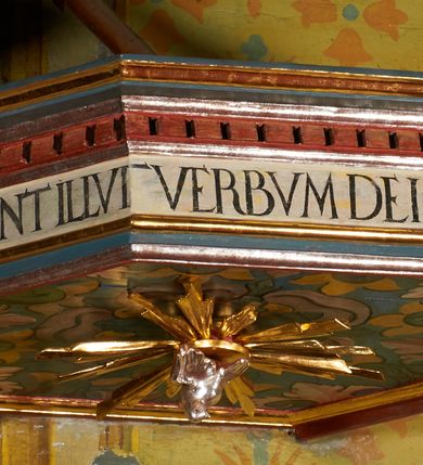 Zdjęcie nr 15: Ambona jest przyścienna, wisząca, z dostępem od zakrystii przez drzwiczki w zaplecku. Kosz ma na planie połowy ośmioboku, na podstawie w formie wałka dekorowanego liśćmi, z podwieszeniem zakończonym szyszką. Ścianki kosza wydzielone są pilastrami rozmieszczonymi w narożach wieloboku, zakończonymi palmetami. Pomiędzy pilastrami znajdują się prostokątne płyciny dekorowane w górnej części ornamentem roślinnym, parapet przyjmuje formę gzymsu. Rolę zaplecka pełnią prostokątne drzwi we wnęce ujęte prostą ramą. Baldachim niepołączony z zapleckiem, na planie ośmioboku, ma formę pełnego belkowania dekorowanego kostkowaniem, w partii fryzu widnieje napis: „A(NNO) D(OMINI) 1601. [CUSTO]DIVNT ILLV[D] VERBVM DEI. BEATI QUI”. Podniebienie baldachimu malowane jest w motywy roślinne z podwieszoną rzeźbioną gołębicą Ducha Świętego w glorii promienistej. Struktura polichromowana jest w kolorze błękitnym, szarym i kremowym, z czerwonymi, pozłacanymi i posrebrzanymi elementami. 

