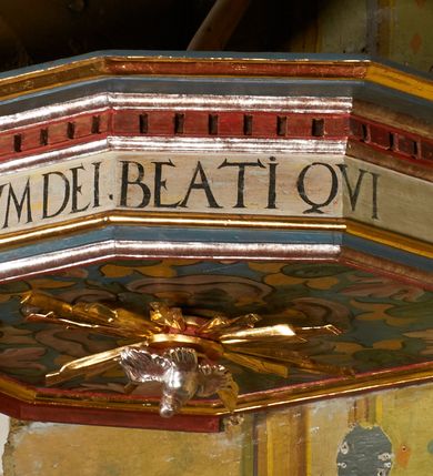 Zdjęcie nr 16: Ambona jest przyścienna, wisząca, z dostępem od zakrystii przez drzwiczki w zaplecku. Kosz ma na planie połowy ośmioboku, na podstawie w formie wałka dekorowanego liśćmi, z podwieszeniem zakończonym szyszką. Ścianki kosza wydzielone są pilastrami rozmieszczonymi w narożach wieloboku, zakończonymi palmetami. Pomiędzy pilastrami znajdują się prostokątne płyciny dekorowane w górnej części ornamentem roślinnym, parapet przyjmuje formę gzymsu. Rolę zaplecka pełnią prostokątne drzwi we wnęce ujęte prostą ramą. Baldachim niepołączony z zapleckiem, na planie ośmioboku, ma formę pełnego belkowania dekorowanego kostkowaniem, w partii fryzu widnieje napis: „A(NNO) D(OMINI) 1601. [CUSTO]DIVNT ILLV[D] VERBVM DEI. BEATI QUI”. Podniebienie baldachimu malowane jest w motywy roślinne z podwieszoną rzeźbioną gołębicą Ducha Świętego w glorii promienistej. Struktura polichromowana jest w kolorze błękitnym, szarym i kremowym, z czerwonymi, pozłacanymi i posrebrzanymi elementami. 

