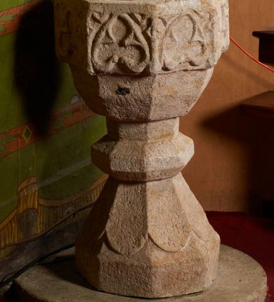 Zdjęcie nr 6: Kamienna chrzcielnica stoi na niewysokiej, kamiennej podstawie na planie koła. Jej stopa jest na planie ośmioboku, u dołu z pionowo ściętym cokołem i zwęża się stożkowato ku górze. Nodus jest spłaszczony, ośmioboczny. Czara ośmioboczna, dwustrefowa, u dołu zaokrąglona, u góry prosto ścięta, nadwieszona, w górnej strefie pola dekorowane są motywem trójlistnego maswerku. Wewnątrz czary znajduje się metalowa misa. Drewniana pokrywa przyjmuje kształt korony zamkniętej sześcioma kabłąkami z kulą i krzyżem umieszczonymi na szczycie. Polichromowana w kolorach zielonym i czerwonym, wewnątrz na niebiesko, obręcz korony i kula posrebrzane, detale i brzegi pozłacane.