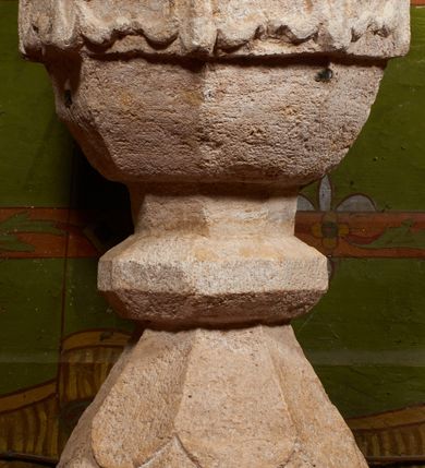 Zdjęcie nr 9: Kamienna chrzcielnica stoi na niewysokiej, kamiennej podstawie na planie koła. Jej stopa jest na planie ośmioboku, u dołu z pionowo ściętym cokołem i zwęża się stożkowato ku górze. Nodus jest spłaszczony, ośmioboczny. Czara ośmioboczna, dwustrefowa, u dołu zaokrąglona, u góry prosto ścięta, nadwieszona, w górnej strefie pola dekorowane są motywem trójlistnego maswerku. Wewnątrz czary znajduje się metalowa misa. Drewniana pokrywa przyjmuje kształt korony zamkniętej sześcioma kabłąkami z kulą i krzyżem umieszczonymi na szczycie. Polichromowana w kolorach zielonym i czerwonym, wewnątrz na niebiesko, obręcz korony i kula posrebrzane, detale i brzegi pozłacane.