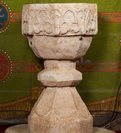 Zdjęcie nr 8: Kamienna chrzcielnica stoi na niewysokiej, kamiennej podstawie na planie koła. Jej stopa jest na planie ośmioboku, u dołu z pionowo ściętym cokołem i zwęża się stożkowato ku górze. Nodus jest spłaszczony, ośmioboczny. Czara ośmioboczna, dwustrefowa, u dołu zaokrąglona, u góry prosto ścięta, nadwieszona, w górnej strefie pola dekorowane są motywem trójlistnego maswerku. Wewnątrz czary znajduje się metalowa misa. Drewniana pokrywa przyjmuje kształt korony zamkniętej sześcioma kabłąkami z kulą i krzyżem umieszczonymi na szczycie. Polichromowana w kolorach zielonym i czerwonym, wewnątrz na niebiesko, obręcz korony i kula posrebrzane, detale i brzegi pozłacane.