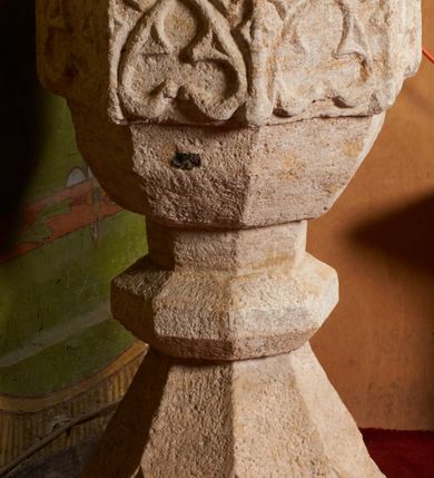 Zdjęcie nr 10: Kamienna chrzcielnica stoi na niewysokiej, kamiennej podstawie na planie koła. Jej stopa jest na planie ośmioboku, u dołu z pionowo ściętym cokołem i zwęża się stożkowato ku górze. Nodus jest spłaszczony, ośmioboczny. Czara ośmioboczna, dwustrefowa, u dołu zaokrąglona, u góry prosto ścięta, nadwieszona, w górnej strefie pola dekorowane są motywem trójlistnego maswerku. Wewnątrz czary znajduje się metalowa misa. Drewniana pokrywa przyjmuje kształt korony zamkniętej sześcioma kabłąkami z kulą i krzyżem umieszczonymi na szczycie. Polichromowana w kolorach zielonym i czerwonym, wewnątrz na niebiesko, obręcz korony i kula posrebrzane, detale i brzegi pozłacane.