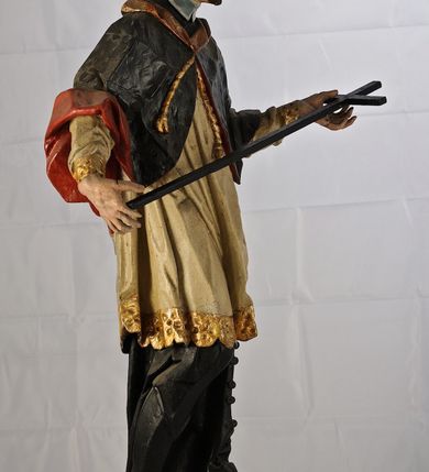 Zdjęcie nr 6: Rzeźba św. Jana Nepomucena jest drążona z tyłu, ustawiona na dwustopniowym cokole, który stanowią skrzynki na relikwie o wyciętych z przodu otworach. Święty ukazany jest w całej postaci, ustawiony frontalnie, w kontrapoście, z pochyloną głową skierowaną w prawo. W obu, rozłożonych na boki rękach podtrzymuje ukośnie ułożony krzyż. Twarz pociągła, o dużym i szerokim nosie, półkoliście zarysowanych łukach brwiowych, małych, migdałowatych oczach oraz pełnych, szerokich ustach. Okolona jest krótką brodą i silnie kręconymi, krótkimi włosami. Święty ubrany jest w zapinaną na guziki czarną sutannę, białą komżę lamowaną płaskorzeźbioną i pozłoconą koronką oraz zawiązaną na piersi i opadającą na ramiona czarną almucję z czerwoną podszewką i pozłacanym kołnierzem (krótką, futrzaną pelerynkę). Figura w partiach ciała polichromowana naturalistycznie. 

