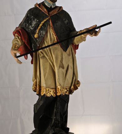 Zdjęcie nr 3: Rzeźba św. Jana Nepomucena jest drążona z tyłu, ustawiona na dwustopniowym cokole, który stanowią skrzynki na relikwie o wyciętych z przodu otworach. Święty ukazany jest w całej postaci, ustawiony frontalnie, w kontrapoście, z pochyloną głową skierowaną w prawo. W obu, rozłożonych na boki rękach podtrzymuje ukośnie ułożony krzyż. Twarz pociągła, o dużym i szerokim nosie, półkoliście zarysowanych łukach brwiowych, małych, migdałowatych oczach oraz pełnych, szerokich ustach. Okolona jest krótką brodą i silnie kręconymi, krótkimi włosami. Święty ubrany jest w zapinaną na guziki czarną sutannę, białą komżę lamowaną płaskorzeźbioną i pozłoconą koronką oraz zawiązaną na piersi i opadającą na ramiona czarną almucję z czerwoną podszewką i pozłacanym kołnierzem (krótką, futrzaną pelerynkę). Figura w partiach ciała polichromowana naturalistycznie. 

