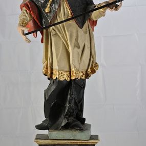 Zdjęcie nr 1: Rzeźba św. Jana Nepomucena jest drążona z tyłu, ustawiona na dwustopniowym cokole, który stanowią skrzynki na relikwie o wyciętych z przodu otworach. Święty ukazany jest w całej postaci, ustawiony frontalnie, w kontrapoście, z pochyloną głową skierowaną w prawo. W obu, rozłożonych na boki rękach podtrzymuje ukośnie ułożony krzyż. Twarz pociągła, o dużym i szerokim nosie, półkoliście zarysowanych łukach brwiowych, małych, migdałowatych oczach oraz pełnych, szerokich ustach. Okolona jest krótką brodą i silnie kręconymi, krótkimi włosami. Święty ubrany jest w zapinaną na guziki czarną sutannę, białą komżę lamowaną płaskorzeźbioną i pozłoconą koronką oraz zawiązaną na piersi i opadającą na ramiona czarną almucję z czerwoną podszewką i pozłacanym kołnierzem (krótką, futrzaną pelerynkę). Figura w partiach ciała polichromowana naturalistycznie. 

