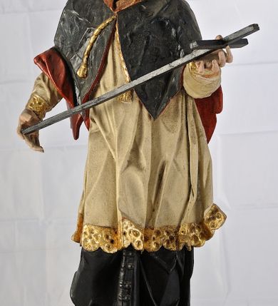 Zdjęcie nr 2: Rzeźba św. Jana Nepomucena jest drążona z tyłu, ustawiona na dwustopniowym cokole, który stanowią skrzynki na relikwie o wyciętych z przodu otworach. Święty ukazany jest w całej postaci, ustawiony frontalnie, w kontrapoście, z pochyloną głową skierowaną w prawo. W obu, rozłożonych na boki rękach podtrzymuje ukośnie ułożony krzyż. Twarz pociągła, o dużym i szerokim nosie, półkoliście zarysowanych łukach brwiowych, małych, migdałowatych oczach oraz pełnych, szerokich ustach. Okolona jest krótką brodą i silnie kręconymi, krótkimi włosami. Święty ubrany jest w zapinaną na guziki czarną sutannę, białą komżę lamowaną płaskorzeźbioną i pozłoconą koronką oraz zawiązaną na piersi i opadającą na ramiona czarną almucję z czerwoną podszewką i pozłacanym kołnierzem (krótką, futrzaną pelerynkę). Figura w partiach ciała polichromowana naturalistycznie. 

