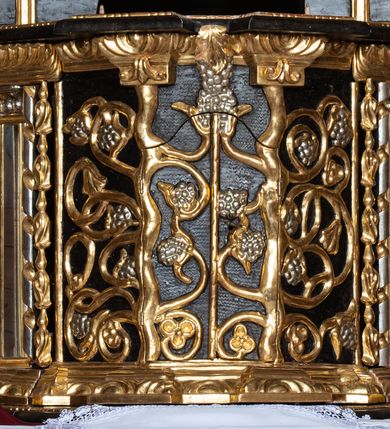 Zdjęcie nr 24: Ołtarz przyścienny, architektoniczny, trójosiowy, dwukondygnacjowy. Nastawa ołtarzowa ustawiona na prostopadłościennym stole ołtarzowym, zasłanym czerwonym materiałem. Predella na niskim cokoliku w formie półwałku, zamknięta u góry puklowanym gzymsem. W osi środkowej wmontowane tabernakulum ozdobione bujną, płaskorzeźbioną winną latoroślą. Po jego bokach ścianki z lustrami, flankowane parą spiralnie skręconych laskowań. W osiach bocznych predelli płyciny o uszatych narożach, w polu których znajdują się pilastry hermowe ze skierowanymi na wprost kobiecymi głowami. Na skrajach predelli konsolki, bogato dekorowane wicią, zawijającą się w wolutki, zdobioną perełkowaniem; na brzegu konsolki skierowana ku dołowi głowa. Pierwsza kondygnacja artykułowana parą kolumn o korynckich kapitelach, wspierających niepełne belkowanie, wyłamane na ich osi. W polu głównym obraz Matki Boskiej z Dzieciątkiem, w osiach bocznych wmontowane skrzydła tryptyku z przedstawieniami: Ecce Homo po lewej stronie oraz Matki Boskiej Bolesnej po prawej stronie. Każda z osi ujęta na brzegach obramowaniem w kształcie stojącego prostokąta o uszatych narożnikach. Fryz dekorowany symetryczną wicią roślinną, nad kolumnami srebrzone rauty. Druga kondygnacja mniejsza, flankowana analogicznymi kolumnami, wspierającymi podobne belkowanie. W polu głównym obraz św. Andrzeja w kształcie stojącego prostokąta. W lewej osi bocznej obraz św. Wojciecha, a w prawej św. Stanisława, oba zamknięte łukiem nadwieszonym. Po bokach kondygnacji uszy o wykroju okuciowym, zdobione wicią oraz podwieszonymi girlandami z owocami winogron. Nad ołtarzem zamocowana promienista gloria, w polu której znajduje się oko Opatrzności otoczone wieńcem z obłoków oraz sześcioma skrzydlatymi główkami anielskimi. Struktura malowana na kolor czarny, kolumny i ornamentyka złocone, detale srebrzone.