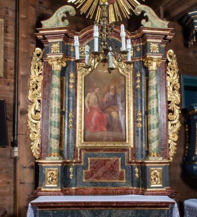 Zdjęcie nr 2: Ołtarz przyścienny, architektoniczny, jednokondygnacyjny, jednoosiowy ze zwieńczeniem. Mensa prostopadłościenna, drewniana, ustawiona na cokole, zamknięta profilowanym gzymsem. Antepedium zdobione malowaną płyciną w kształcie leżącego prostokąta o wklęsłych narożach i zryzalitowanym górnym i dolnym boku. W polu baranek apokaliptyczny na księdze zamkniętej siedmioma pieczęciami, po bokach girlandy kwiatowe, tło ciemnozielone. Nastawa ołtarzowa ustawiona na niskim cokole o wysuniętych do przodu impostach, na których ustawiona jest para kolumn, wspierających przerwane belkowanie i wolutowy przyczółek. Pośrodku w ramie ozdobionej płaskorzeźbionymi liśćmi obraz Trójcy Świętej w kształcie stojącego prostokąta, zamkniętego łukiem wklęsło-wypukłym z uskokami. Po bokach obrazu zwisy z camapnul. Fryz dekorowany rozetkami i liśćmi akantu. Za kolumnami prostokątne płyciny, marmoryzowane. W zwieńczeniu płaskorzeźba św. Tekli z krzyżem i płonącym stosem na tle obłoków i promienistej glorii z czterema uskrzydlonymi główkami anielskimi. Po bokach ołtarza akantowe uszy, przeplecione karbowaną wstęgą. Imposty w partii cokołu zdobione płycinami w kształcie niewielkich prostokątów, wypełnione liśćmi akantu. Struktura marmoryzowana na kolor ciemnoniebieski, trzony kolumn marmoryzowane na kolor zielono-beżowy, profilowania na kolor bordowy, detale i ornamentyka złocone. 