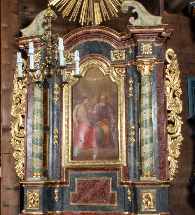 Zdjęcie nr 1: Ołtarz przyścienny, architektoniczny, jednokondygnacyjny, jednoosiowy ze zwieńczeniem. Mensa prostopadłościenna, drewniana, ustawiona na cokole, zamknięta profilowanym gzymsem. Antepedium zdobione malowaną płyciną w kształcie leżącego prostokąta o wklęsłych narożach i zryzalitowanym górnym i dolnym boku. W polu baranek apokaliptyczny na księdze zamkniętej siedmioma pieczęciami, po bokach girlandy kwiatowe, tło ciemnozielone. Nastawa ołtarzowa ustawiona na niskim cokole o wysuniętych do przodu impostach, na których ustawiona jest para kolumn, wspierających przerwane belkowanie i wolutowy przyczółek. Pośrodku w ramie ozdobionej płaskorzeźbionymi liśćmi obraz Trójcy Świętej w kształcie stojącego prostokąta, zamkniętego łukiem wklęsło-wypukłym z uskokami. Po bokach obrazu zwisy z camapnul. Fryz dekorowany rozetkami i liśćmi akantu. Za kolumnami prostokątne płyciny, marmoryzowane. W zwieńczeniu płaskorzeźba św. Tekli z krzyżem i płonącym stosem na tle obłoków i promienistej glorii z czterema uskrzydlonymi główkami anielskimi. Po bokach ołtarza akantowe uszy, przeplecione karbowaną wstęgą. Imposty w partii cokołu zdobione płycinami w kształcie niewielkich prostokątów, wypełnione liśćmi akantu. Struktura marmoryzowana na kolor ciemnoniebieski, trzony kolumn marmoryzowane na kolor zielono-beżowy, profilowania na kolor bordowy, detale i ornamentyka złocone. 