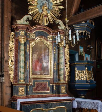 Zdjęcie nr 3: Ołtarz przyścienny, architektoniczny, jednokondygnacyjny, jednoosiowy ze zwieńczeniem. Mensa prostopadłościenna, drewniana, ustawiona na cokole, zamknięta profilowanym gzymsem. Antepedium zdobione malowaną płyciną w kształcie leżącego prostokąta o wklęsłych narożach i zryzalitowanym górnym i dolnym boku. W polu baranek apokaliptyczny na księdze zamkniętej siedmioma pieczęciami, po bokach girlandy kwiatowe, tło ciemnozielone. Nastawa ołtarzowa ustawiona na niskim cokole o wysuniętych do przodu impostach, na których ustawiona jest para kolumn, wspierających przerwane belkowanie i wolutowy przyczółek. Pośrodku w ramie ozdobionej płaskorzeźbionymi liśćmi obraz Trójcy Świętej w kształcie stojącego prostokąta, zamkniętego łukiem wklęsło-wypukłym z uskokami. Po bokach obrazu zwisy z camapnul. Fryz dekorowany rozetkami i liśćmi akantu. Za kolumnami prostokątne płyciny, marmoryzowane. W zwieńczeniu płaskorzeźba św. Tekli z krzyżem i płonącym stosem na tle obłoków i promienistej glorii z czterema uskrzydlonymi główkami anielskimi. Po bokach ołtarza akantowe uszy, przeplecione karbowaną wstęgą. Imposty w partii cokołu zdobione płycinami w kształcie niewielkich prostokątów, wypełnione liśćmi akantu. Struktura marmoryzowana na kolor ciemnoniebieski, trzony kolumn marmoryzowane na kolor zielono-beżowy, profilowania na kolor bordowy, detale i ornamentyka złocone. 