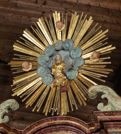 Zdjęcie nr 4: Ołtarz przyścienny, architektoniczny, jednokondygnacyjny, jednoosiowy ze zwieńczeniem. Mensa prostopadłościenna, drewniana, ustawiona na cokole, zamknięta profilowanym gzymsem. Antepedium zdobione malowaną płyciną w kształcie leżącego prostokąta o wklęsłych narożach i zryzalitowanym górnym i dolnym boku. W polu baranek apokaliptyczny na księdze zamkniętej siedmioma pieczęciami, po bokach girlandy kwiatowe, tło ciemnozielone. Nastawa ołtarzowa ustawiona na niskim cokole o wysuniętych do przodu impostach, na których ustawiona jest para kolumn, wspierających przerwane belkowanie i wolutowy przyczółek. Pośrodku w ramie ozdobionej płaskorzeźbionymi liśćmi obraz Trójcy Świętej w kształcie stojącego prostokąta, zamkniętego łukiem wklęsło-wypukłym z uskokami. Po bokach obrazu zwisy z camapnul. Fryz dekorowany rozetkami i liśćmi akantu. Za kolumnami prostokątne płyciny, marmoryzowane. W zwieńczeniu płaskorzeźba św. Tekli z krzyżem i płonącym stosem na tle obłoków i promienistej glorii z czterema uskrzydlonymi główkami anielskimi. Po bokach ołtarza akantowe uszy, przeplecione karbowaną wstęgą. Imposty w partii cokołu zdobione płycinami w kształcie niewielkich prostokątów, wypełnione liśćmi akantu. Struktura marmoryzowana na kolor ciemnoniebieski, trzony kolumn marmoryzowane na kolor zielono-beżowy, profilowania na kolor bordowy, detale i ornamentyka złocone. 