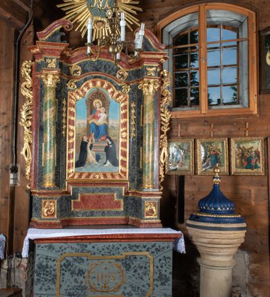 Zdjęcie nr 5: Ołtarz przyścienny, architektoniczny, jednokondygnacyjny, jednoosiowy ze zwieńczeniem. Mensa prostopadłościenna, drewniana, ustawiona na cokole, zamknięta profilowanym gzymsem. Antepedium zdobione malowaną płyciną w kształcie leżącego prostokąta o wklęsłych narożach i zryzalitowanym górnym i dolnym boku. W polu monogram IHS otoczony wieńcem z kłosów zbóż. Tło jasnoniebieskie, fakturowane w ciemnoszare plamki. Nastawa ołtarzowa ustawiona na niskim cokole o wysuniętych do przodu impostach, na których ustawiona jest para kolumn, wspierających przerwane belkowanie i wolutowy przyczółek. Pośrodku w ramie ozdobionej płaskorzeźbionymi liśćmi obraz Matki Boskiej Różańcowej w kształcie stojącego prostokąta, zamkniętego łukiem wklęsło-wypukłym z uskokami. Po bokach obrazu zwisy z kamapnul. Fryz dekorowany liśćmi akantu i rozetkami. Na tle kolumn prostokątne płyciny. W zwieńczeniu monogram maryjny na tle promienistej glorii z czterema uskrzydlonymi główkami anielskimi. Po bokach ołtarza akantowe uszy, przeplecione karbowaną wstęgą. Imposty w partii cokołu zdobione płycinami w kształcie niewielkich prostokątów, wypełnionych liśćmi akantu. Struktura malowana na kolor ciemnoniebieski i fakturowana w szare plamki, trzony kolumn marmoryzowane na kolor zielono-beżowy, profilowania na kolor bordowy, detale i ornamentyka złocone. 