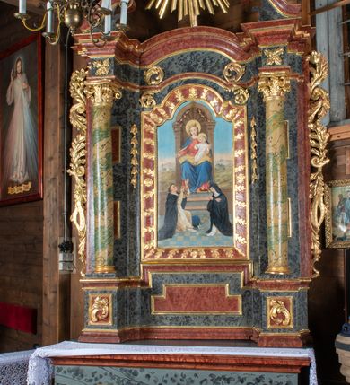 Zdjęcie nr 3: Ołtarz przyścienny, architektoniczny, jednokondygnacyjny, jednoosiowy ze zwieńczeniem. Mensa prostopadłościenna, drewniana, ustawiona na cokole, zamknięta profilowanym gzymsem. Antepedium zdobione malowaną płyciną w kształcie leżącego prostokąta o wklęsłych narożach i zryzalitowanym górnym i dolnym boku. W polu monogram IHS otoczony wieńcem z kłosów zbóż. Tło jasnoniebieskie, fakturowane w ciemnoszare plamki. Nastawa ołtarzowa ustawiona na niskim cokole o wysuniętych do przodu impostach, na których ustawiona jest para kolumn, wspierających przerwane belkowanie i wolutowy przyczółek. Pośrodku w ramie ozdobionej płaskorzeźbionymi liśćmi obraz Matki Boskiej Różańcowej w kształcie stojącego prostokąta, zamkniętego łukiem wklęsło-wypukłym z uskokami. Po bokach obrazu zwisy z kamapnul. Fryz dekorowany liśćmi akantu i rozetkami. Na tle kolumn prostokątne płyciny. W zwieńczeniu monogram maryjny na tle promienistej glorii z czterema uskrzydlonymi główkami anielskimi. Po bokach ołtarza akantowe uszy, przeplecione karbowaną wstęgą. Imposty w partii cokołu zdobione płycinami w kształcie niewielkich prostokątów, wypełnionych liśćmi akantu. Struktura malowana na kolor ciemnoniebieski i fakturowana w szare plamki, trzony kolumn marmoryzowane na kolor zielono-beżowy, profilowania na kolor bordowy, detale i ornamentyka złocone. 