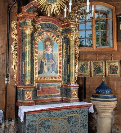 Zdjęcie nr 4: Ołtarz przyścienny, architektoniczny, jednokondygnacyjny, jednoosiowy ze zwieńczeniem. Mensa prostopadłościenna, drewniana, ustawiona na cokole, zamknięta profilowanym gzymsem. Antepedium zdobione malowaną płyciną w kształcie leżącego prostokąta o wklęsłych narożach i zryzalitowanym górnym i dolnym boku. W polu monogram IHS otoczony wieńcem z kłosów zbóż. Tło jasnoniebieskie, fakturowane w ciemnoszare plamki. Nastawa ołtarzowa ustawiona na niskim cokole o wysuniętych do przodu impostach, na których ustawiona jest para kolumn, wspierających przerwane belkowanie i wolutowy przyczółek. Pośrodku w ramie ozdobionej płaskorzeźbionymi liśćmi obraz Matki Boskiej Różańcowej w kształcie stojącego prostokąta, zamkniętego łukiem wklęsło-wypukłym z uskokami. Po bokach obrazu zwisy z kamapnul. Fryz dekorowany liśćmi akantu i rozetkami. Na tle kolumn prostokątne płyciny. W zwieńczeniu monogram maryjny na tle promienistej glorii z czterema uskrzydlonymi główkami anielskimi. Po bokach ołtarza akantowe uszy, przeplecione karbowaną wstęgą. Imposty w partii cokołu zdobione płycinami w kształcie niewielkich prostokątów, wypełnionych liśćmi akantu. Struktura malowana na kolor ciemnoniebieski i fakturowana w szare plamki, trzony kolumn marmoryzowane na kolor zielono-beżowy, profilowania na kolor bordowy, detale i ornamentyka złocone. 