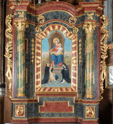 Zdjęcie nr 1: Ołtarz przyścienny, architektoniczny, jednokondygnacyjny, jednoosiowy ze zwieńczeniem. Mensa prostopadłościenna, drewniana, ustawiona na cokole, zamknięta profilowanym gzymsem. Antepedium zdobione malowaną płyciną w kształcie leżącego prostokąta o wklęsłych narożach i zryzalitowanym górnym i dolnym boku. W polu monogram IHS otoczony wieńcem z kłosów zbóż. Tło jasnoniebieskie, fakturowane w ciemnoszare plamki. Nastawa ołtarzowa ustawiona na niskim cokole o wysuniętych do przodu impostach, na których ustawiona jest para kolumn, wspierających przerwane belkowanie i wolutowy przyczółek. Pośrodku w ramie ozdobionej płaskorzeźbionymi liśćmi obraz Matki Boskiej Różańcowej w kształcie stojącego prostokąta, zamkniętego łukiem wklęsło-wypukłym z uskokami. Po bokach obrazu zwisy z kamapnul. Fryz dekorowany liśćmi akantu i rozetkami. Na tle kolumn prostokątne płyciny. W zwieńczeniu monogram maryjny na tle promienistej glorii z czterema uskrzydlonymi główkami anielskimi. Po bokach ołtarza akantowe uszy, przeplecione karbowaną wstęgą. Imposty w partii cokołu zdobione płycinami w kształcie niewielkich prostokątów, wypełnionych liśćmi akantu. Struktura malowana na kolor ciemnoniebieski i fakturowana w szare plamki, trzony kolumn marmoryzowane na kolor zielono-beżowy, profilowania na kolor bordowy, detale i ornamentyka złocone. 